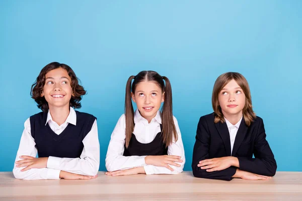 Foto de colegiales bonitos de ensueño usan brazos de mesa sentados uniformes plegados buscando espacio vacío sonriendo aislado color azul fondo — Foto de Stock