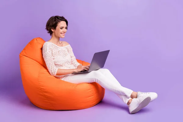 Foto penuh wanita menawan yang ceria duduk dengan kursi empuk menggunakan laptop yang terisolasi pada latar belakang warna ungu — Stok Foto