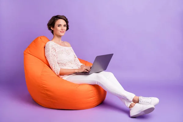Foto wanita muda yang menawan manis mengenakan kemeja rajutan duduk beanbag mengetik gadget modern terisolasi latar belakang warna ungu — Stok Foto