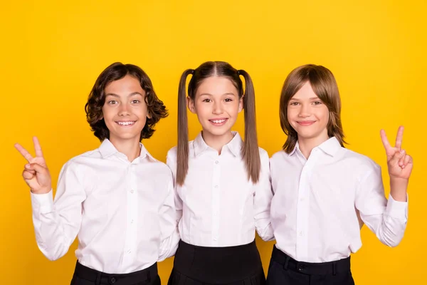 Foto de tres amigables escolares muestran gesto v-signo desgaste camisa blanca aislado color amarillo fondo —  Fotos de Stock