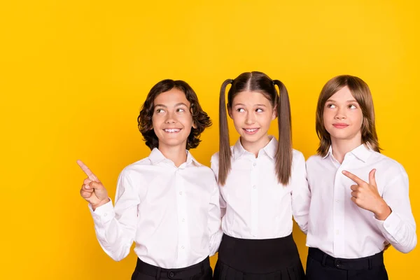 Photo of three schoolkids direct finger empty space look up wear white shirt isolated yellow color background — Stock Photo, Image