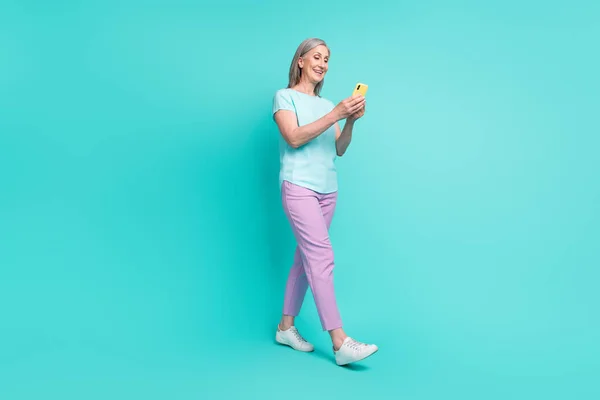 원문 기사보기 전반적 인 크기의 웃고 있는 성숙 한 여성 이 청록색 배경에서 고립된 스마트폰에 중독된 채걸어 들어가는 모습 — 스톡 사진
