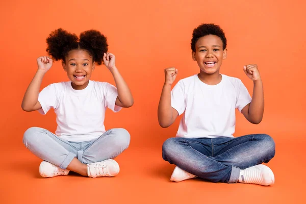 Foto de tamaño completo de poco hooray niña niño sentarse manos puños gritar usar blanco camiseta jeans zapatillas aisladas sobre fondo naranja —  Fotos de Stock