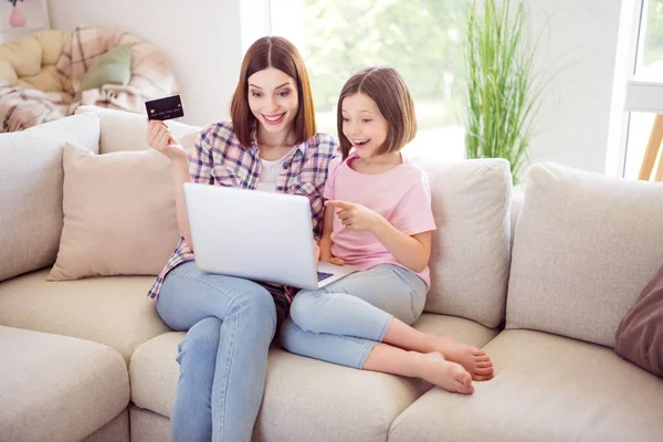 Фото счастливых людей брюнетки сестры сидеть диван смотреть ноутбук указывать пальцем держать кредитную карту в помещении дома — стоковое фото