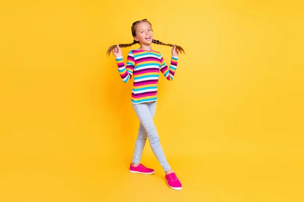 Photo of excited funny little lady play braids enjoy walk wear striped shirt jeans shoes isolated yellow color background — Stock Photo, Image