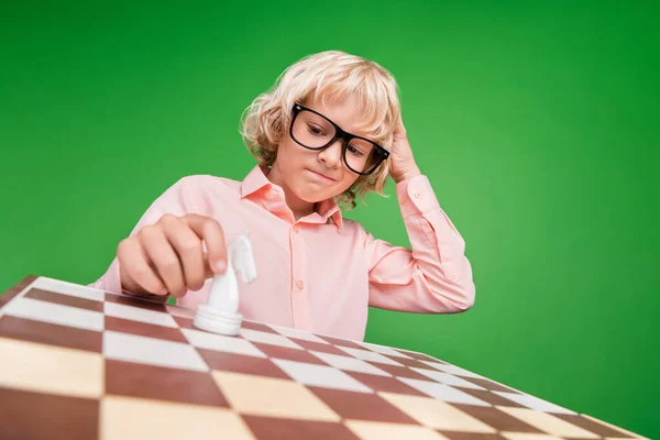 Foto de un joven chico desconcertado mira el tablero piensa mover el caballo de ajedrez aislado sobre fondo de color verde —  Fotos de Stock