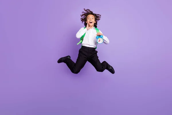 Фотографія повного розміру вражений маленький брюнетка хлопчик стрибок тримати годинник носити сумку сорочки штани кросівки ізольовані на фіолетовому фоні — стокове фото