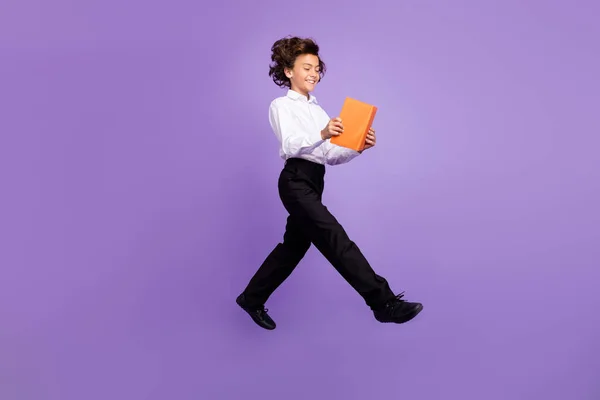 Plná délka fotografie funky malý brunet chlapec skok držet knihu nosit košile kalhoty izolované na fialové barvy pozadí — Stock fotografie