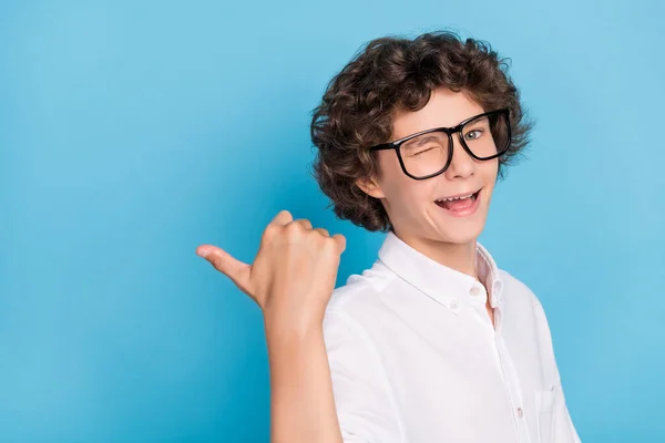 Foto de amigável engraçado escola menino desgaste branco camisa óculos piscando apontando polegar vazio espaço sorrindo isolado azul cor fundo — Fotografia de Stock