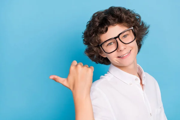 Foto de encantador bonito estudante desgaste branco camisa óculos sorrindo apontando polegar de volta vazio espaço isolado azul cor fundo — Fotografia de Stock