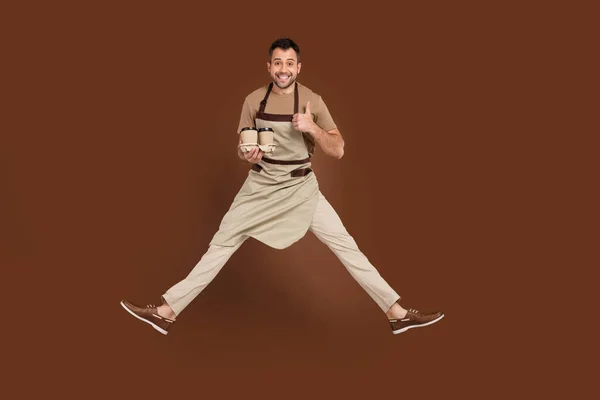 Foto de confiado joven dulce vestido beige uniforme saltando mostrando el pulgar hacia arriba preparando latte aislado color marrón fondo — Foto de Stock