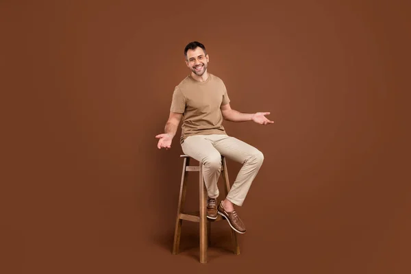 Foto retrato morena homem em pastel t-shirt sorrindo sentado na cadeira de madeira isolado cor marrom fundo — Fotografia de Stock