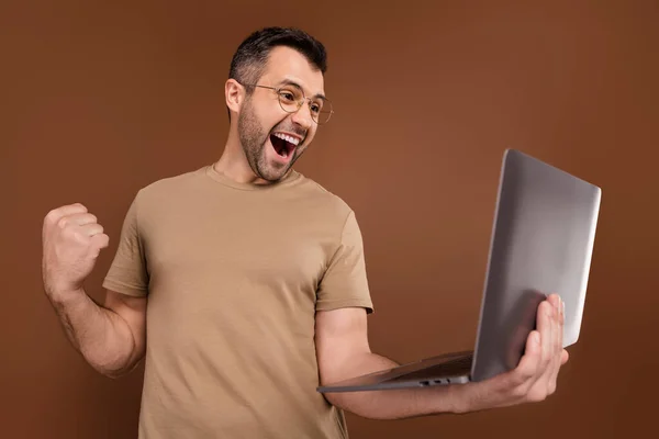 Foto portret brunet man in pastel t-shirt houden laptop gebaren als winnaar geïsoleerde bruine kleur achtergrond — Stockfoto