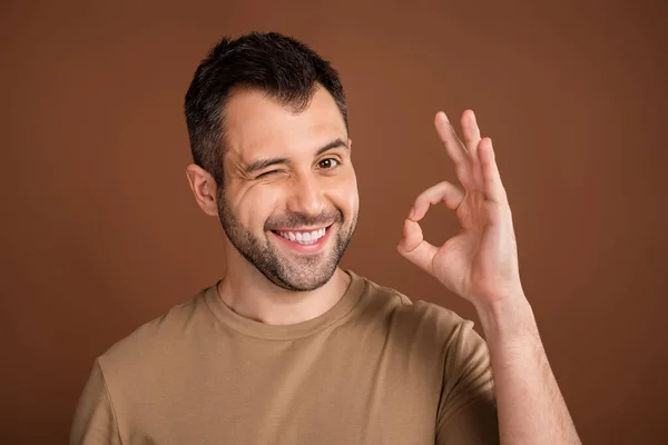 Foto av rolig söt ung kille klädd beige kläder ler blinkande visar okey isolerad brun färg bakgrund — Stockfoto