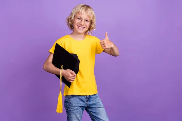 Foto de menino bonito atraente escola desgaste amarelo t-shirt óculos segurando argamassa mostrando polegar para cima isolado cor roxa fundo — Fotografia de Stock