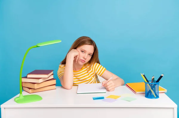 Fénykép elégedetlen iskolás tartsa toll ül az asztal mögött néz kamera elszigetelt kék színű háttér — Stock Fotó