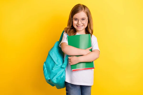 Фото очаровательной веселой школьницы носить белые футболки рюкзак улыбаясь обнимая копировальные книги изолированный желтый цвет фона — стоковое фото