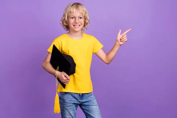 Photo de garçon diplômé tenir chapeau montrent espace vide porter jaune t-shirt isolé fond de couleur pourpre — Photo