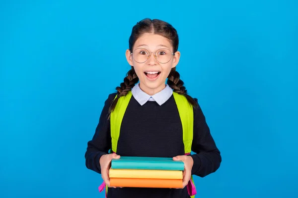 Foto de alegre emocionado colegiala loca en gafas celebrar libros de pila preparar tarea aislado sobre fondo de color azul —  Fotos de Stock