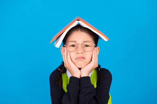 Photo of unhappy upset tired schoolgirl in glasses prepare exam hold book head isolated on blue color background — Stock Photo, Image