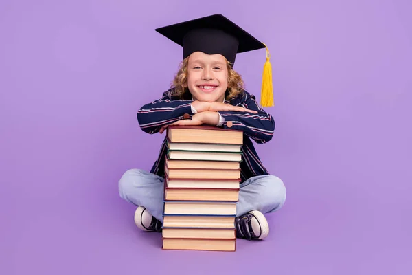 Ritratto di bel scolaro allegro che indossa cappello seduto con libro pila isolato su sfondo viola viola — Foto Stock