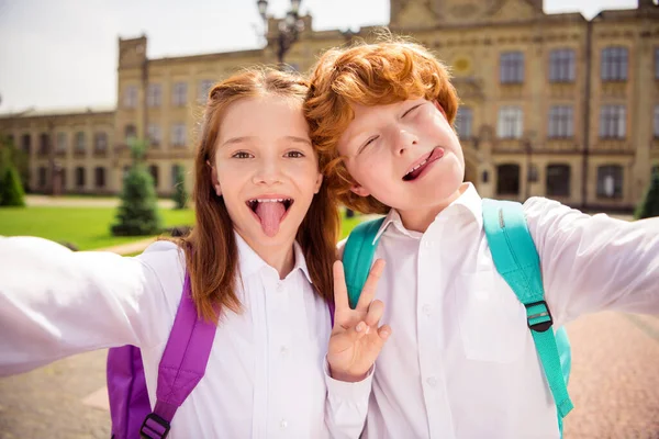 Foto de dois estudantes adoráveis funky fazer selfie mostrar língua v-sinal desgaste saco branco camisa uniforme parque fora — Fotografia de Stock