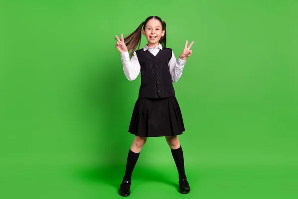 Fénykép fényes bájos iskolás lány viselni fekete fehér egyenruha mutatja v-jelek mosolygó elszigetelt zöld szín háttér — Stock Fotó
