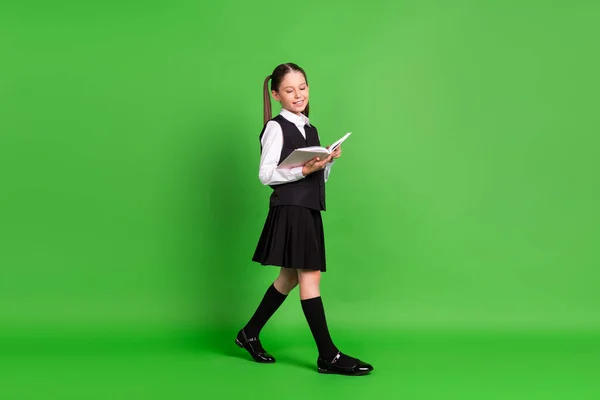 Foto av bedårande söt skolflicka klädd vit svart uniform ler läsa bok promenader isolerad grön färg bakgrund — Stockfoto