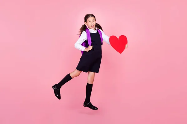 Full längd kropp storlek foto skolflicka hoppa med ryggsäck hålla rött hjärta isolerad pastell rosa färg bakgrund — Stockfoto