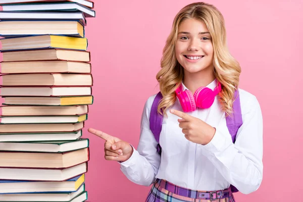 Foto van schattig zoet schoolmeisje dragen formele outfit rugzak hoofdtelefoon wijzend boek stack glimlachen geïsoleerde roze kleur achtergrond — Stockfoto