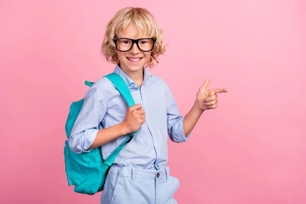 Foto de doce adorável escola menino desgaste azul camisa óculos mochila apontando dedo vazio espaço sorrindo isolado cor-de-rosa fundo — Fotografia de Stock