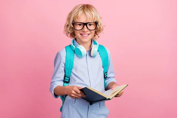 Foto di dolce funky ragazzo della scuola indossare occhiali camicia blu zaino auricolari sorridente lettura libro isolato colore rosa sfondo — Foto Stock