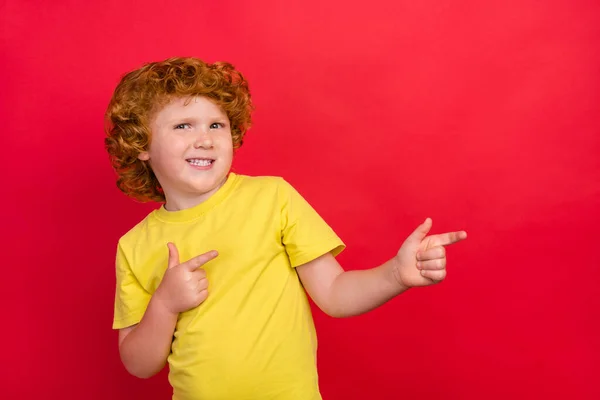 Foto av ingefära liten pojke indikerar pekfingrar tomt utrymme bära gul t-shirt isolerad röd färg bakgrund — Stockfoto