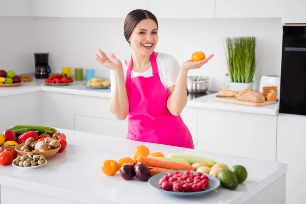 Φωτογραφία από τα εντυπωσιακά καστανά μαλλιά νεαρή κοπέλα κρατήσει ντομάτα από το σπίτι φορούν λευκό t-shirt ποδιά στην κουζίνα — Φωτογραφία Αρχείου