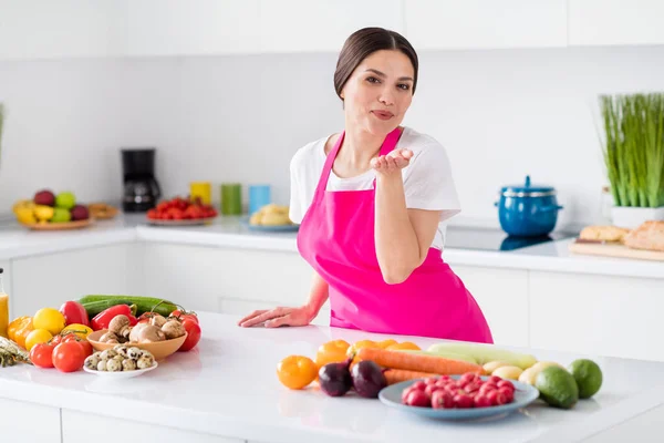 Φωτογραφία του φλερτ γοητευτική ώριμη γυναίκα ντυμένος ροζ ποδιά στέλνοντας σας φιλί μαγείρεμα μεσημεριανό γεύμα σε εσωτερικούς χώρους σπίτι δωμάτιο — Φωτογραφία Αρχείου