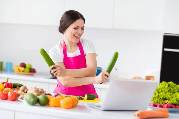 Tatlı, komik, genç bir bayanın pembe önlük giyip, elinde salatalıkla yemek pişirirken, evde gülümserken fotoğrafı. — Stok fotoğraf