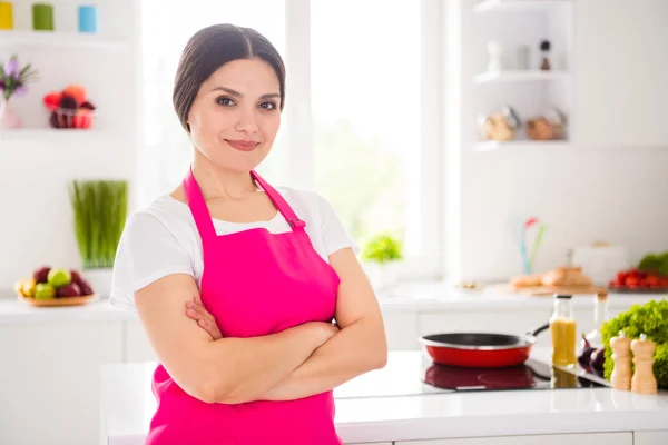 Фотопортрет жінки в рожевому фартусі, посміхаючись зі складеними руками на кухні перед приготуванням вечері — стокове фото