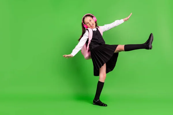 Foto de adorable niña de la escuela dulce usar negro blanco uniforme mochila auriculares caminando sonriendo aislado color verde fondo —  Fotos de Stock