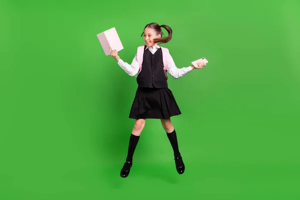 Çok tatlı bir kızın fotoğrafı siyah beyaz bir üniforma giyiyor. Sırt çantasından atlıyor. Elinde modern bir aletle yeşil bir arka plan tutuyor. — Stok fotoğraf