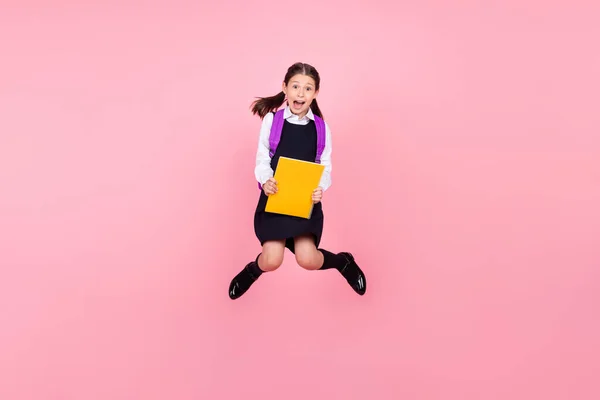 Full längd kropp storlek foto skolflicka hoppa med skolväska hålla bok förvånad isolerad pastell rosa färg bakgrund — Stockfoto