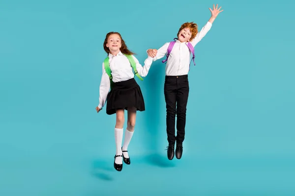 Foto av två funky drömmande barn hoppa gå skolan bära ryggsäck vit skjorta uniform isolerad blå färg bakgrund — Stockfoto