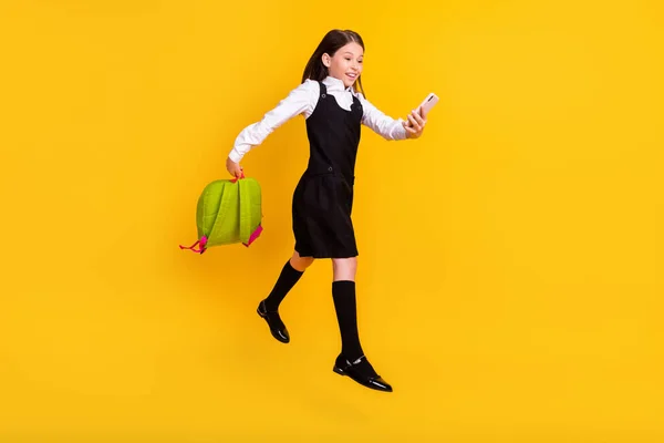 Longitud completa cuerpo tamaño foto colegiala saltar con mochila usando teléfono inteligente aislado vibrante color amarillo fondo —  Fotos de Stock