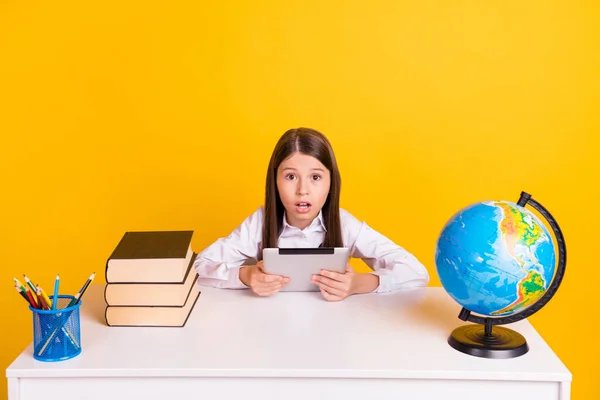 Foto porträtt skolflicka sitter vid skrivbordet förvånad hålla e-läsare i skolan isolerad levande gul färg bakgrund — Stockfoto