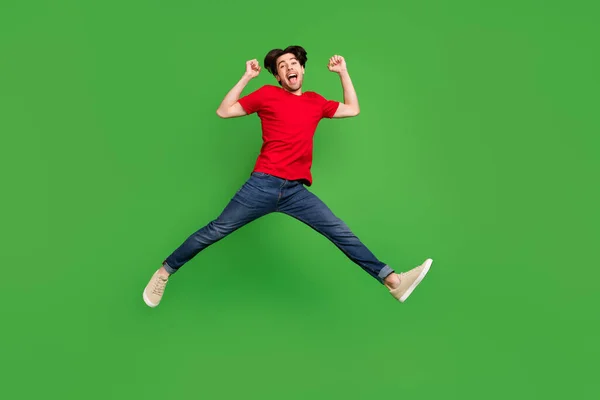 Foto van gekke actieve sportieve kerel springen vieren overwinning dragen rode t-shirt jeans schoenen geïsoleerde groene kleur achtergrond — Stockfoto