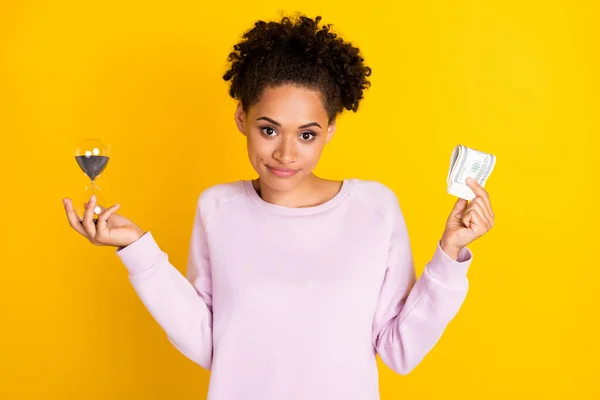 Portret van positieve mooie donkere huid dame houden geld zandglas kijken camera geïsoleerd op gele kleur achtergrond — Stockfoto