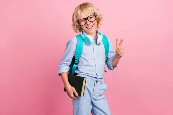 Foto van vrij aantrekkelijke schooljongen dragen bril rugzak hoofdtelefoon v-teken holding boek geïsoleerde roze kleur achtergrond — Stockfoto