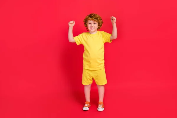 Foto de alegre ganador niño pequeño levante los puños celebrar la victoria usar camiseta amarilla aislado fondo de color rojo — Foto de Stock