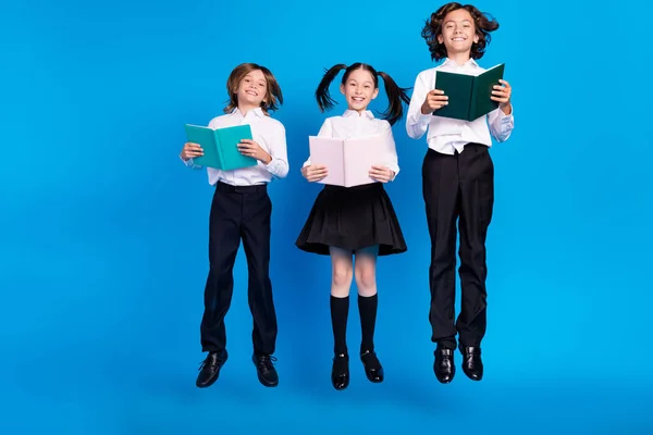 Фото беззаботных школьников прыгать читать домашнюю литературу носить однородную обувь изолированный синий цвет фона — стоковое фото