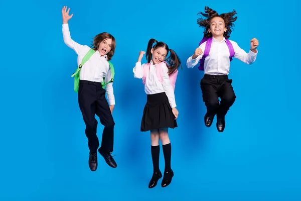 Bild av imponerade förvånade skolbarn bära enhetliga ryggsäckar stigande knytnävar hoppa högt leende isolerad blå färg bakgrund — Stockfoto