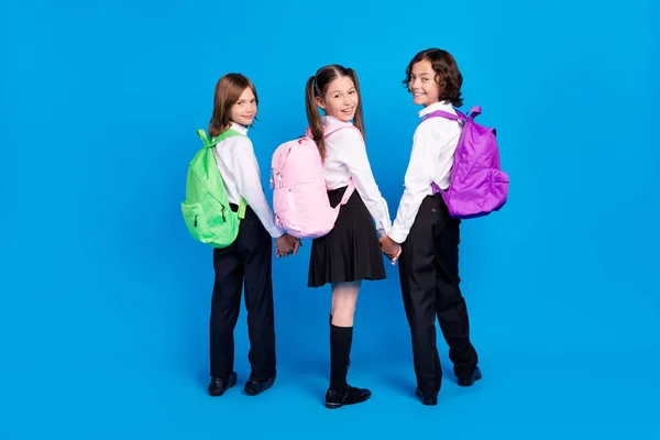 Foto de escolares muito adoráveis usam mochilas uniformes segurando braços andando sorrindo isolado fundo de cor azul — Fotografia de Stock
